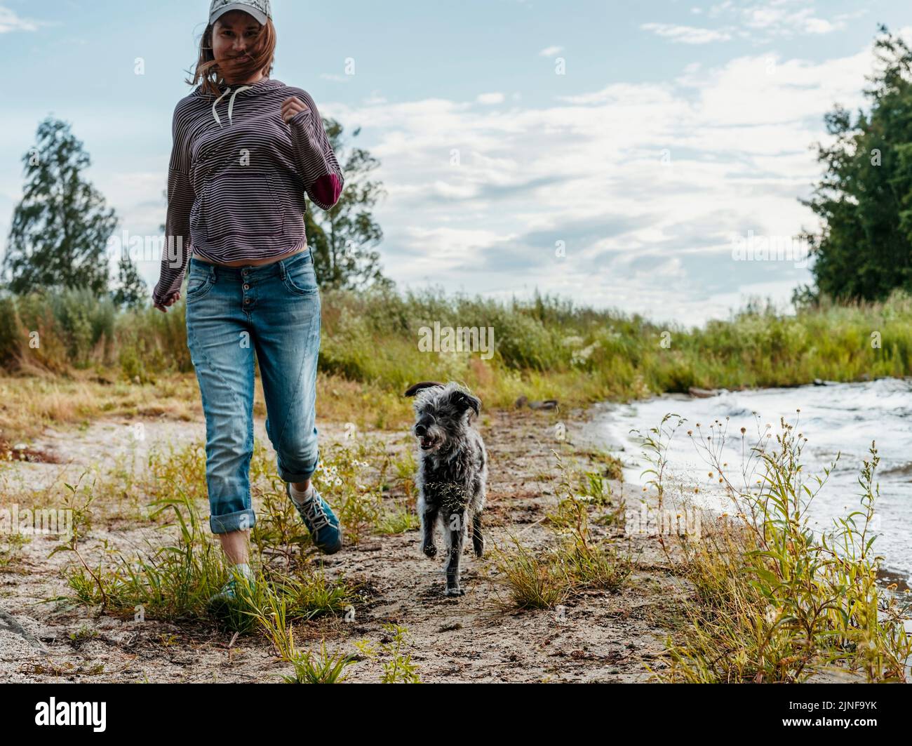 feliz joven mujer corriendo a lo largo de la orilla del lago con la mezcla de razas de perros de viaje y senderismo con mascotas perro caminar Foto de stock