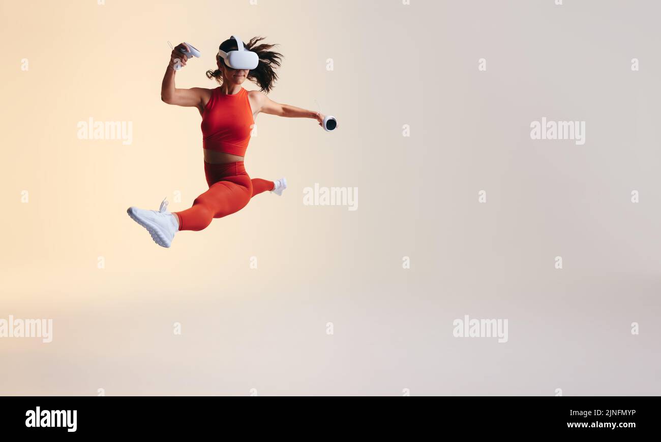 Mujer joven atlética sosteniendo los mandos de juego mientras corría en el aire. Mujer joven deportiva teniendo una sesión de entrenamiento en realidad virtual. Jóvenes activos w Foto de stock
