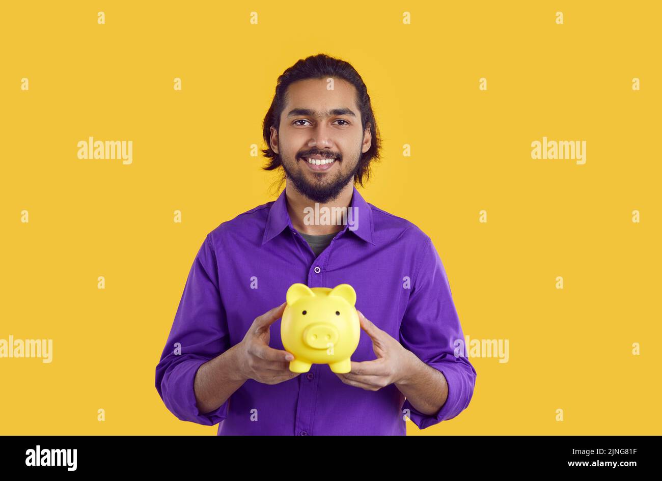 Joven hombre indio feliz con sonrisa muestra una banco piggy simbolizando el ahorro de dinero se encuentra en el estudio Foto de stock