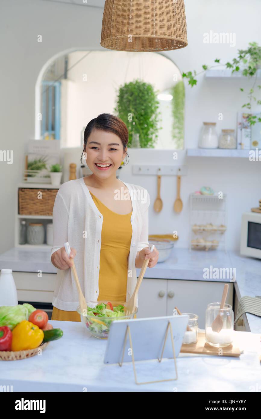 Mujer joven viendo la receta en una tableta digital mientras cocina el almuerzo en la cocina Foto de stock