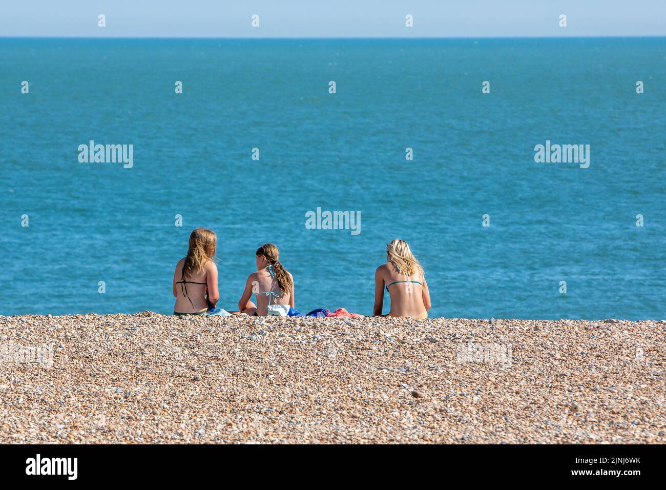 Tres mujeres jóvenes tomando el sol en una playa de guijarros junto a un mar azul y tranquilo Foto de stock