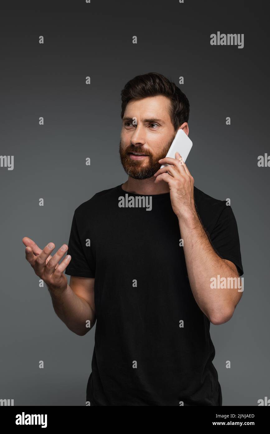 hombre barbudo en camiseta negra hablando en smartphone aislado sobre imagen de stock gris Foto de stock