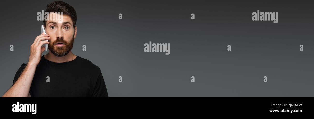 hombre sorprendido en negro camiseta hablando en smartphone aislado en gris, banner, imagen de archivo Foto de stock