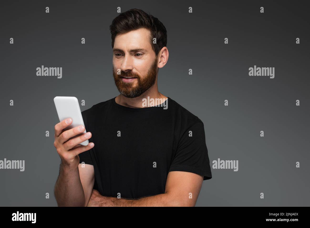un hombre barbudo sonriente en una camiseta negra mirando un smartphone aislado sobre una imagen de stock gris Foto de stock