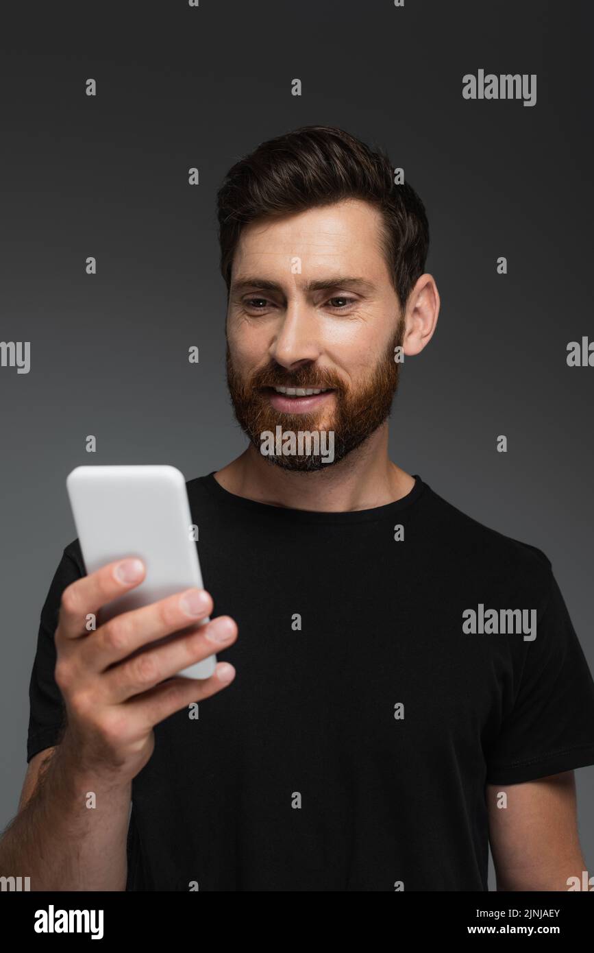 hombre contento y barbudo en negro camiseta con teléfono móvil aislado en gris, imagen de stock Foto de stock