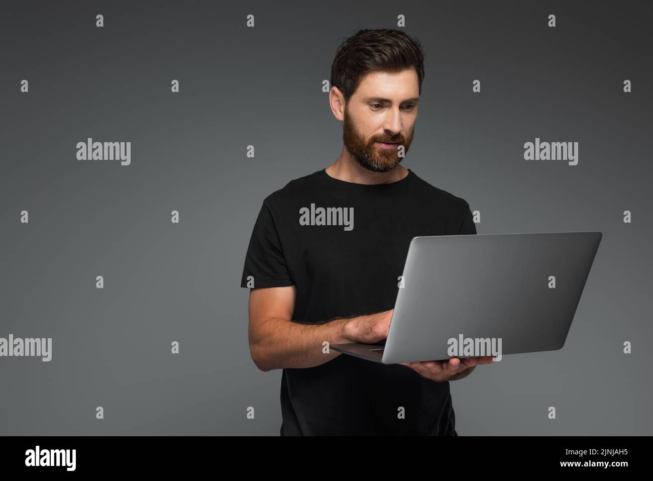 hombre barbudo en camiseta negra con portátil aislado sobre imagen de stock gris Foto de stock