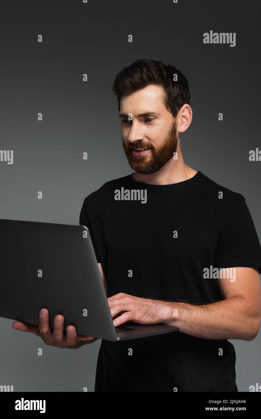 hombre alegre y barbudo en camiseta negra con portátil aislado sobre imagen de stock gris Foto de stock