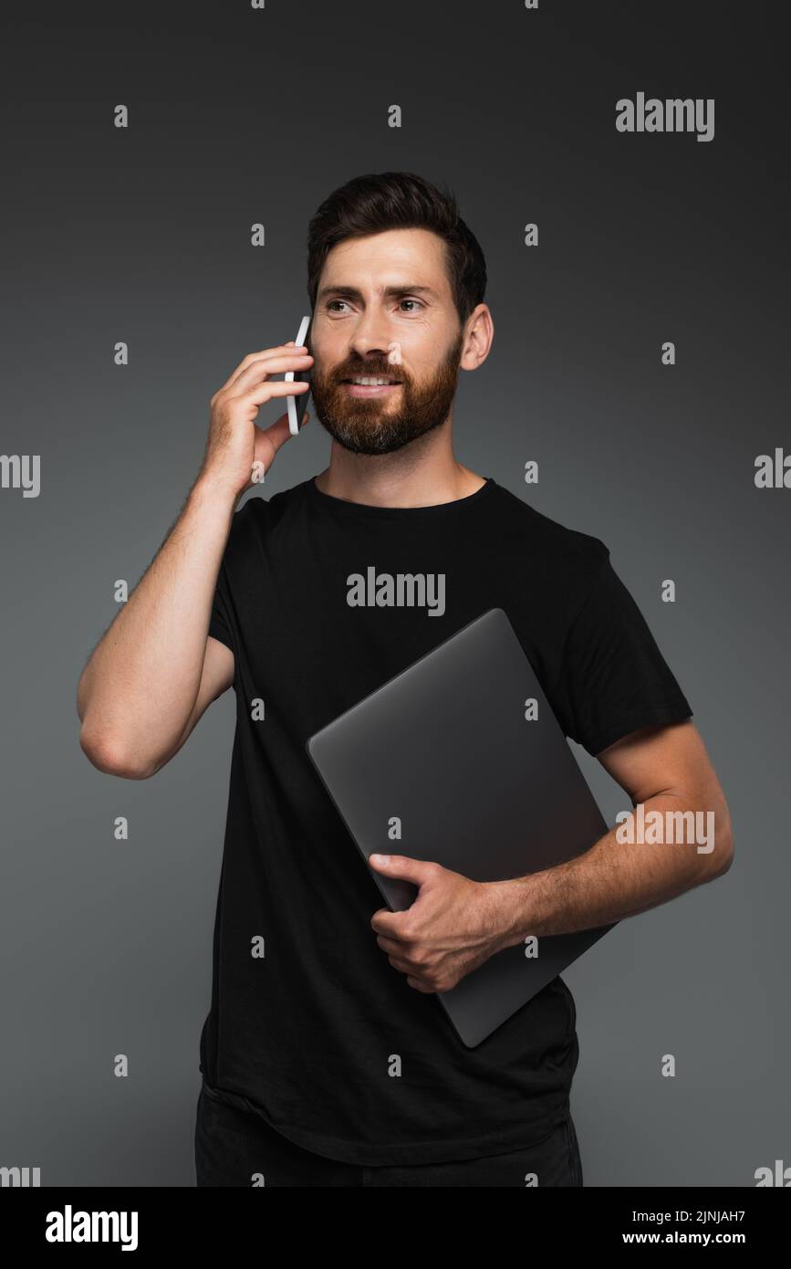 feliz hombre barbudo en camiseta negra sosteniendo el portátil mientras habla en smartphone aislado en gris, imagen de archivo Foto de stock