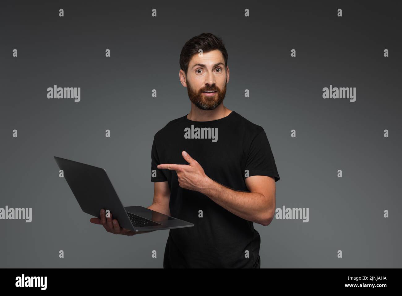 hombre asombrado y barbudo en camiseta negra apuntando con el dedo al ordenador portátil aislado sobre imagen de archivo gris Foto de stock