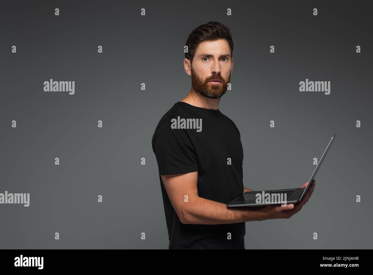 barbudo freelancer en negro camiseta portátil de sujeción aislado en gris, imagen de stock Foto de stock