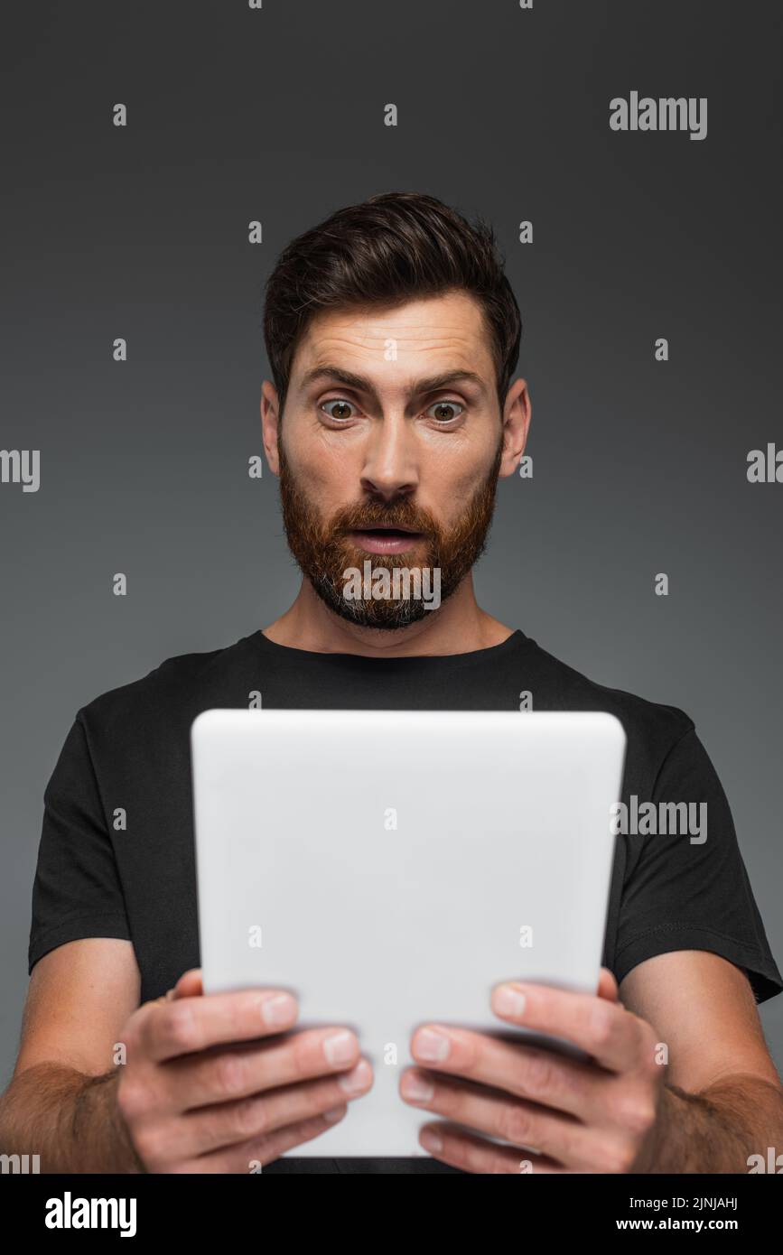 hombre sorprendido y barbudo en negro camiseta sosteniendo la tableta digital aislado en gris, imagen de archivo Foto de stock