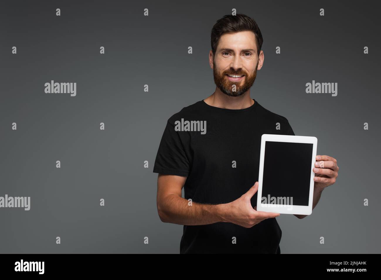 feliz hombre barbudo en negro camiseta sosteniendo tableta digital con pantalla en blanco aislado en gris, imagen de stock Foto de stock