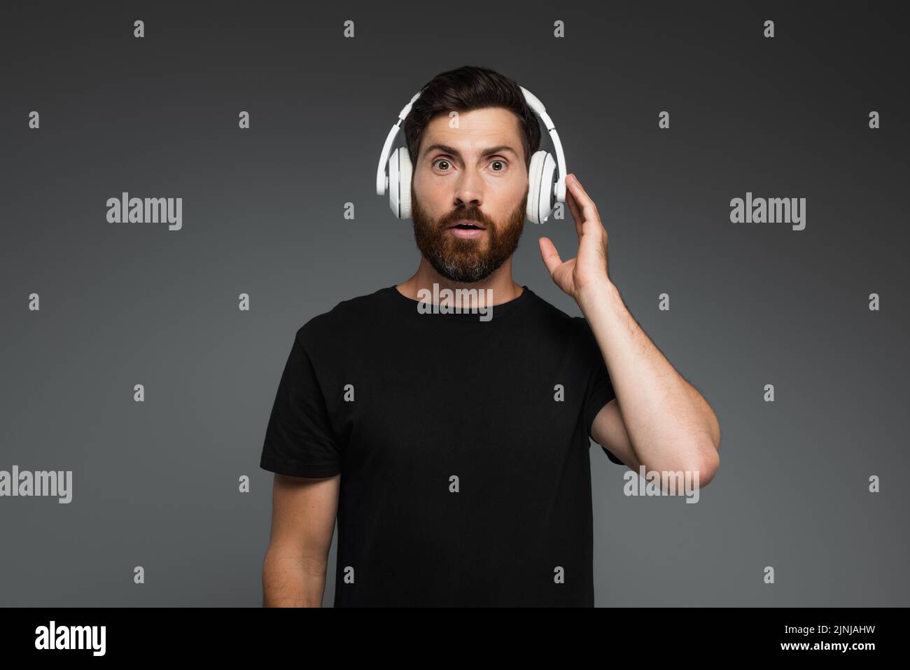 sorprendido hombre tocando los auriculares inalámbricos y escuchando música aislada en gris, imagen de archivo Foto de stock