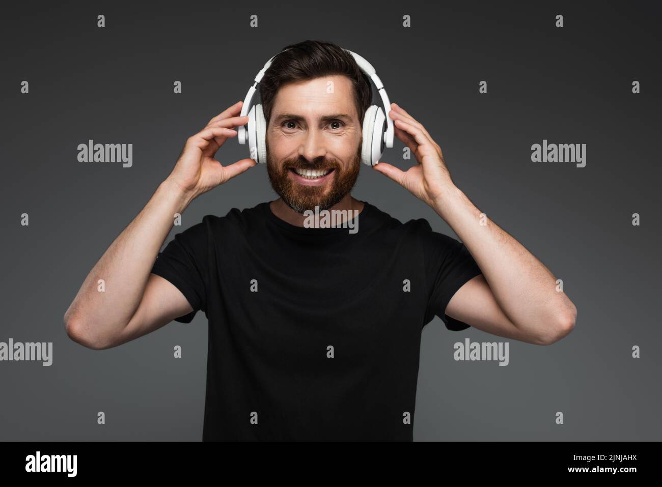 un hombre alegre y barbudo con auriculares inalámbricos escuchando música aislada sobre una imagen de stock gris Foto de stock