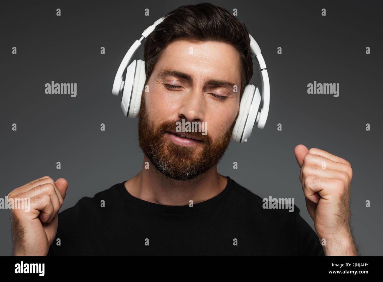 hombre barbudo con los ojos cerrados escuchando música en auriculares inalámbricos aislados en gris, imagen de stock Foto de stock