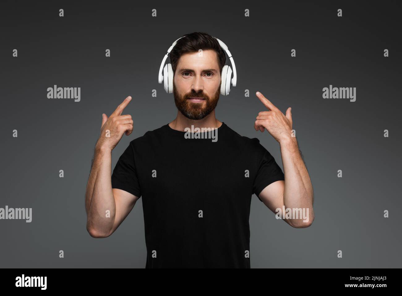 hombre barbudo escuchando música y apuntando con los dedos a los auriculares inalámbricos aislados en una imagen de stock gris Foto de stock