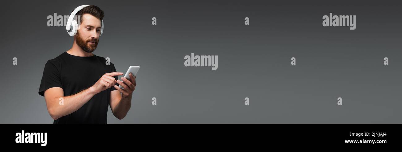 hombre barbudo con auriculares inalámbricos escuchando música y apuntando al smartphone aislado en gris, banner, imagen de stock Foto de stock