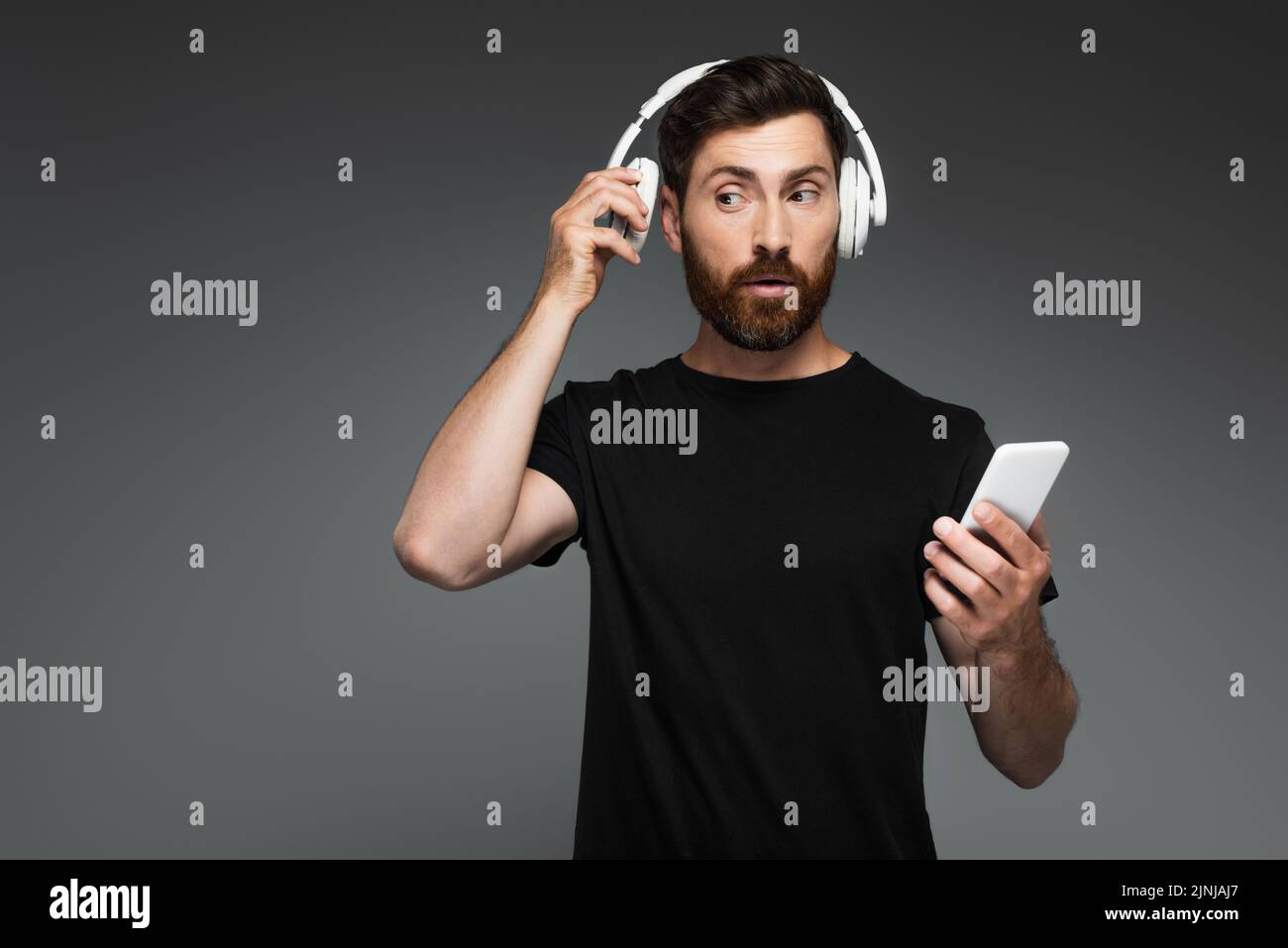 hombre curioso que ajusta los auriculares inalámbricos y utiliza el smartphone aislado sobre una imagen de stock gris Foto de stock