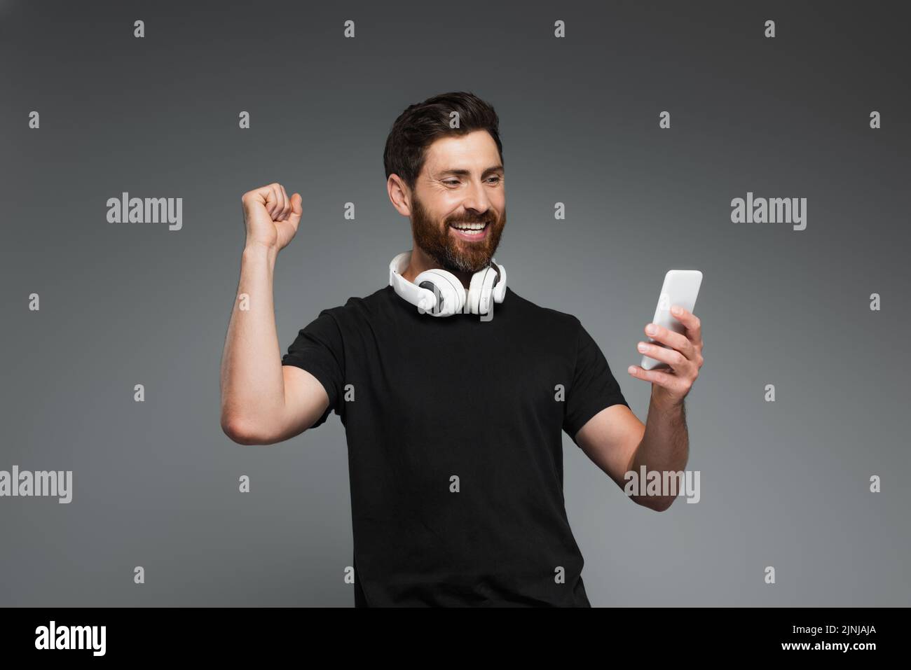hombre barbudo emocionado con auriculares inalámbricos con smartphone aislado sobre imagen de stock gris Foto de stock