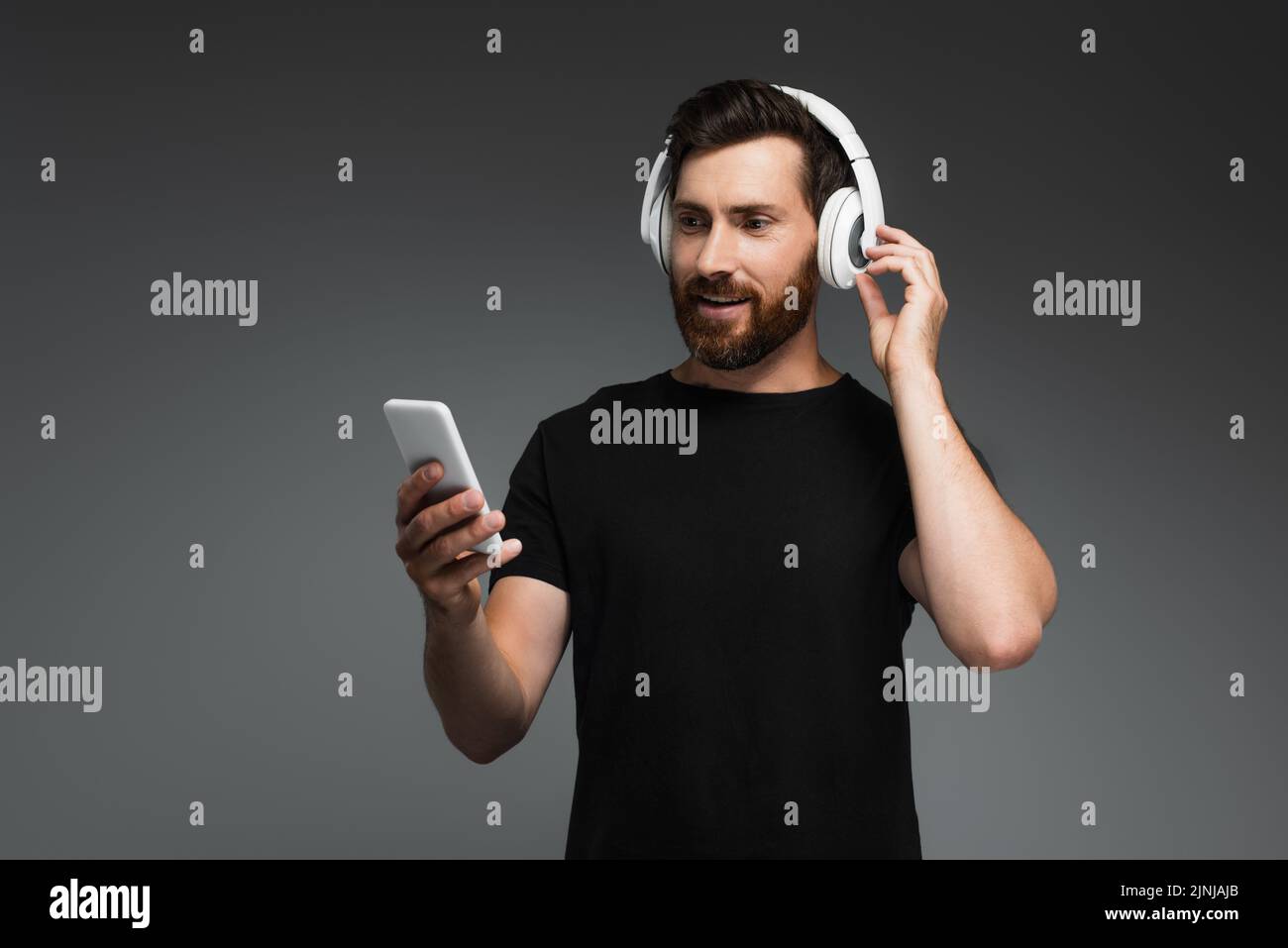 hombre sonriendo con auriculares inalámbricos escuchando música y utilizando un smartphone aislado sobre una imagen de stock gris Foto de stock