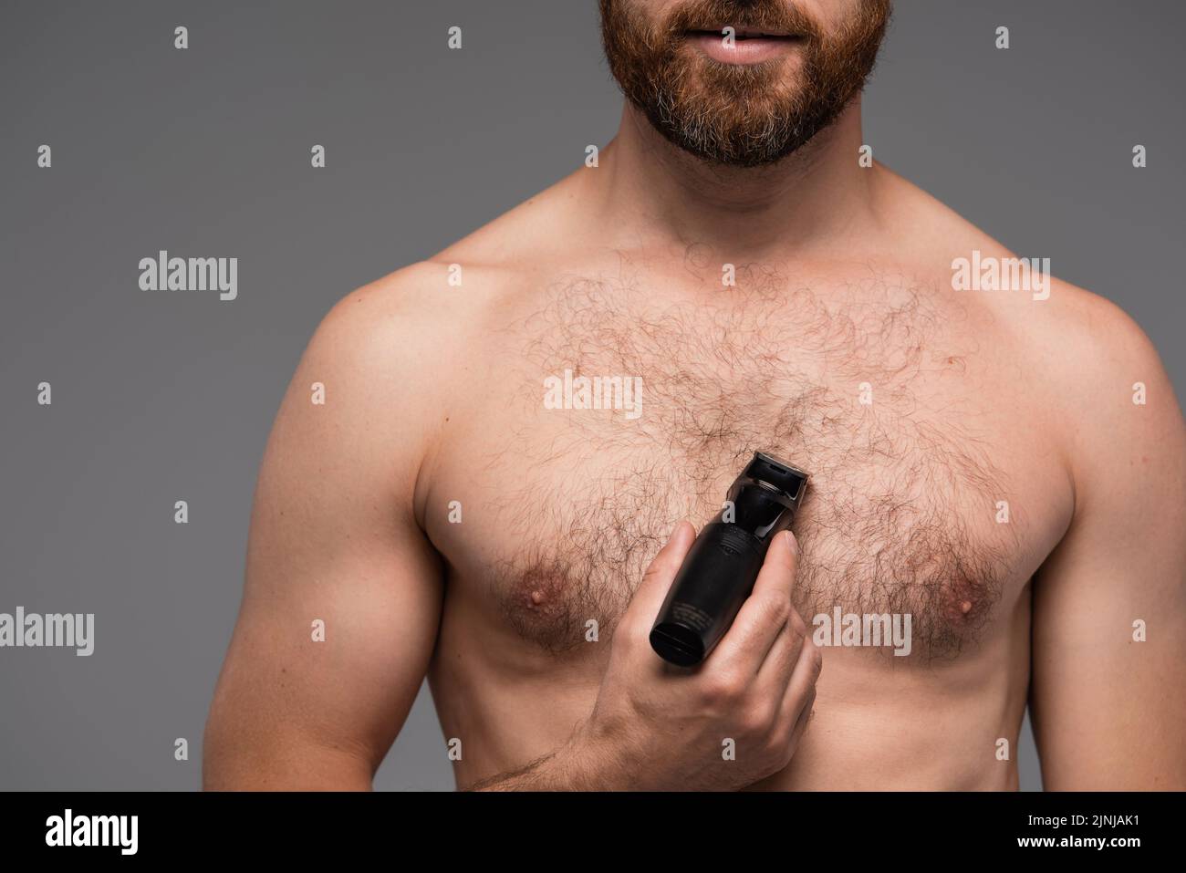 vista recortada de hombre sin camisa afeitándose el pelo en el pecho con afeitadora eléctrica aislada sobre imagen de archivo gris Foto de stock