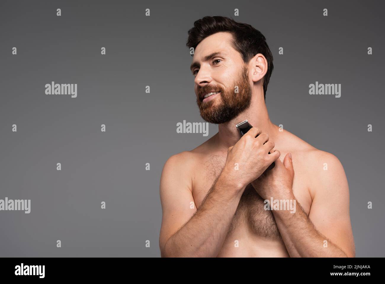 hombre alegre y sin camisa con pelo en el pecho afeitándose con afeitadora eléctrica aislada en gris, imagen de archivo Foto de stock