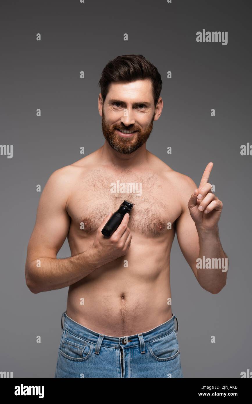 hombre feliz y sin camisa afeitarse el pelo en el pecho con la afeitadora eléctrica y gesturing aislado en gris, imagen de archivo Foto de stock