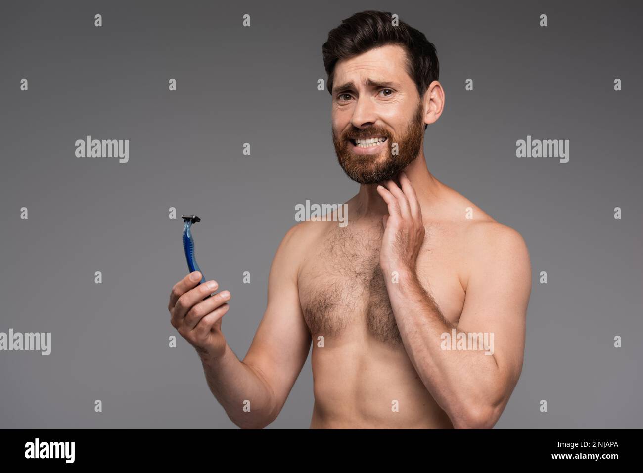 hombre disgustado y sin camisa con la barba sujetando la afeitadora de seguridad aislada sobre la imagen de archivo gris Foto de stock