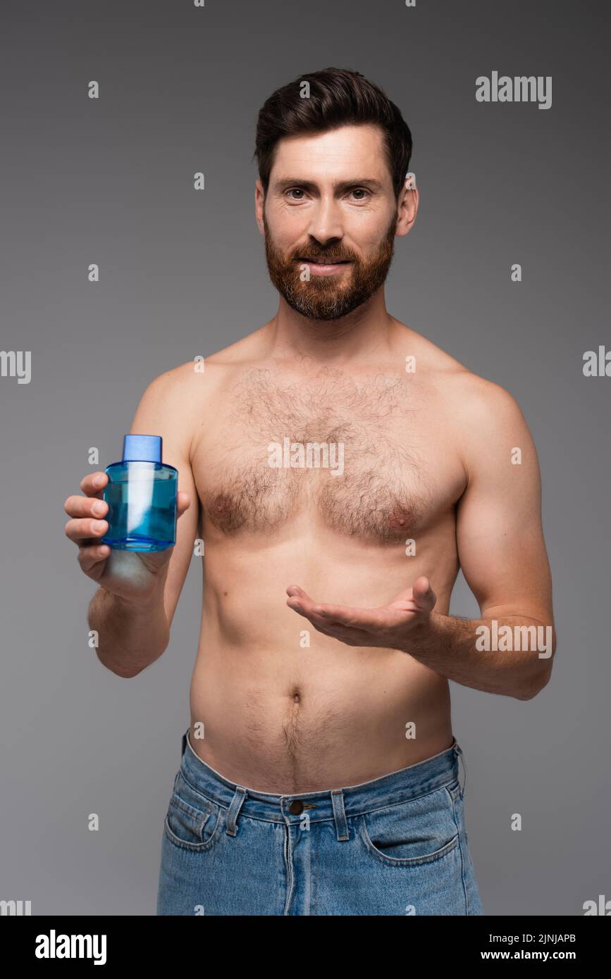 hombre barbudo y sin camisa apuntando con la mano a la botella con producto after shave aislado sobre imagen de stock gris Foto de stock