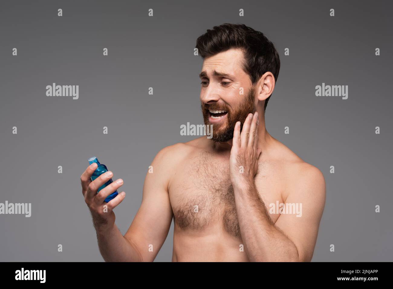 botella de sujeción de hombre con barba y sin camisa con producto after shave aislada sobre imagen de stock gris Foto de stock