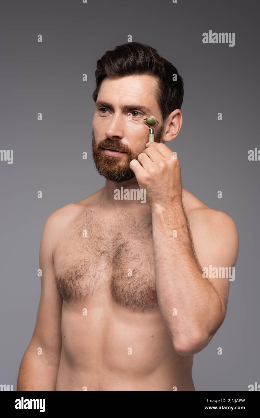 hombre sin camisa con barba utilizando un rodillo de jade mientras masajea la cara aislada sobre una imagen de stock gris Foto de stock