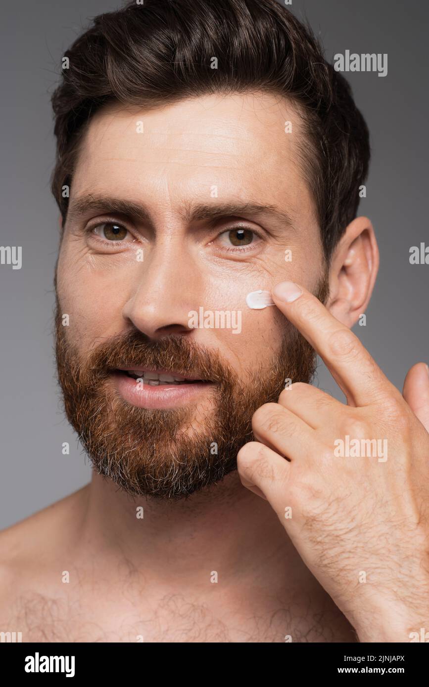 primer plano del hombre con la barba aplicando crema sobre la cara aislada sobre imagen de archivo gris Foto de stock