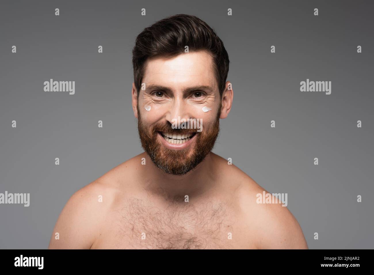 hombre alegre y barbudo con crema en la cara sonriendo aislado en gris, imagen de archivo Foto de stock
