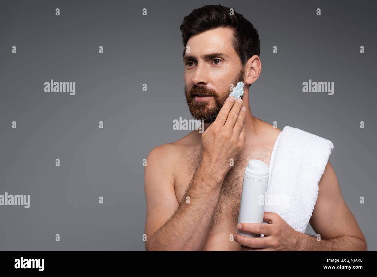 hombre barbado con toalla aplicando espuma de afeitar aislada sobre imagen de stock gris Foto de stock