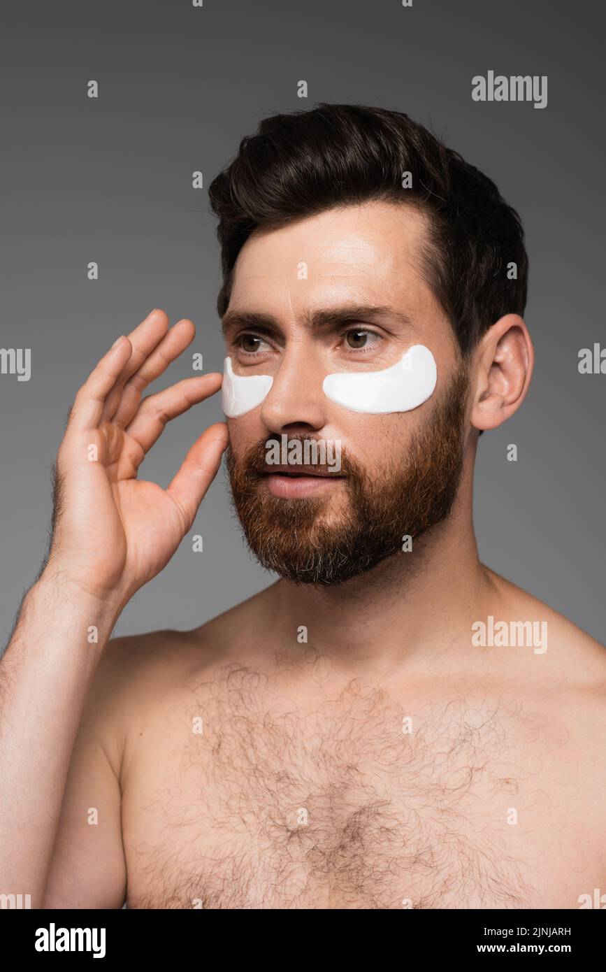hombre barbudo aplicando parches oculares hidratantes aislados sobre imagen de stock gris Foto de stock
