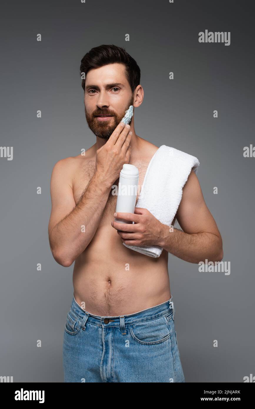 hombre barbudo con toalla aplicando espuma de afeitar y mirando la cámara aislada en gris, imagen de stock Foto de stock
