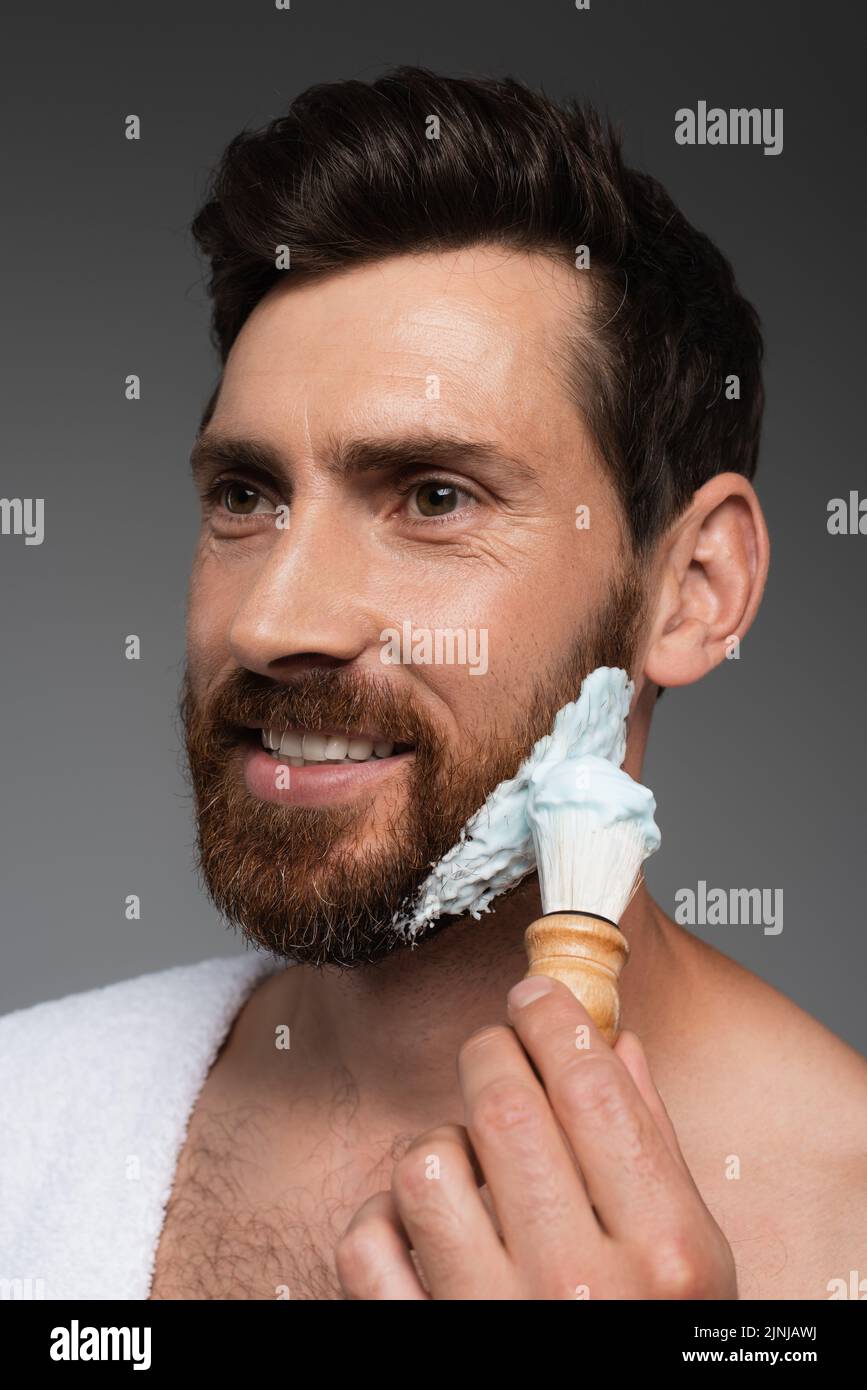 retrato de hombre barbudo feliz aplicando espuma de afeitar con cepillo de afeitar aislado sobre imagen de stock gris Foto de stock