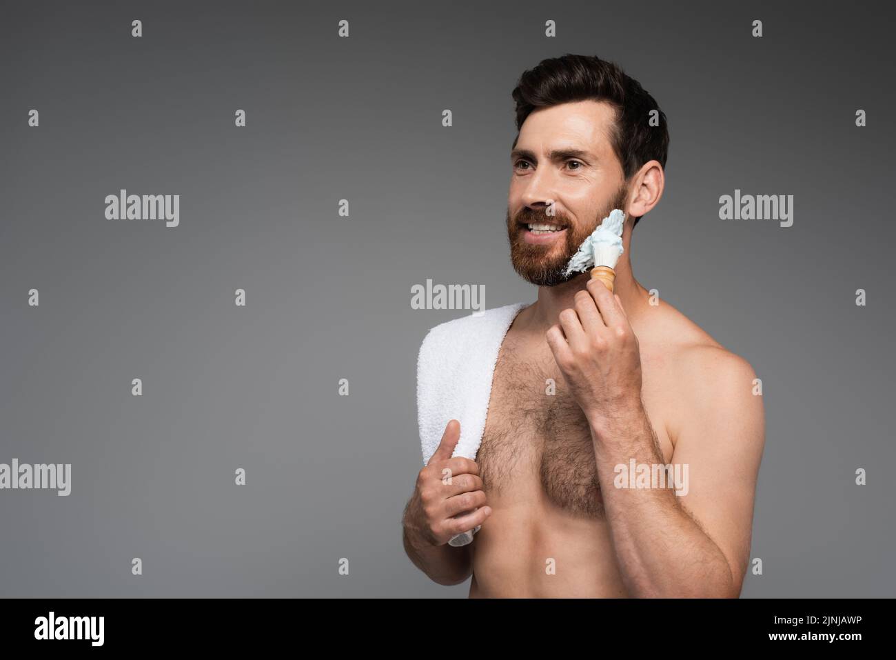 hombre barbudo feliz con toalla aplicando espuma de afeitado con cepillo de afeitado aislado sobre imagen de stock gris Foto de stock