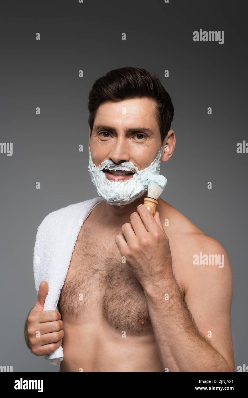 hombre alegre aplicando espuma de afeitar blanca en la cara y sonriendo aislado en gris, imagen de archivo Foto de stock