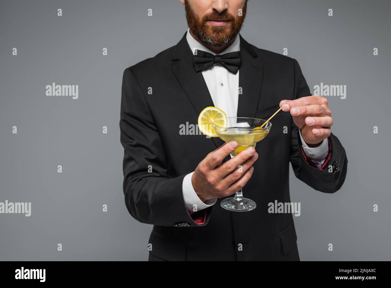 vista parcial de hombre barbudo en traje con pajarita sujetando palillo de dientes con oliva en vidrio con martini aislado en gris, imagen de stock Foto de stock