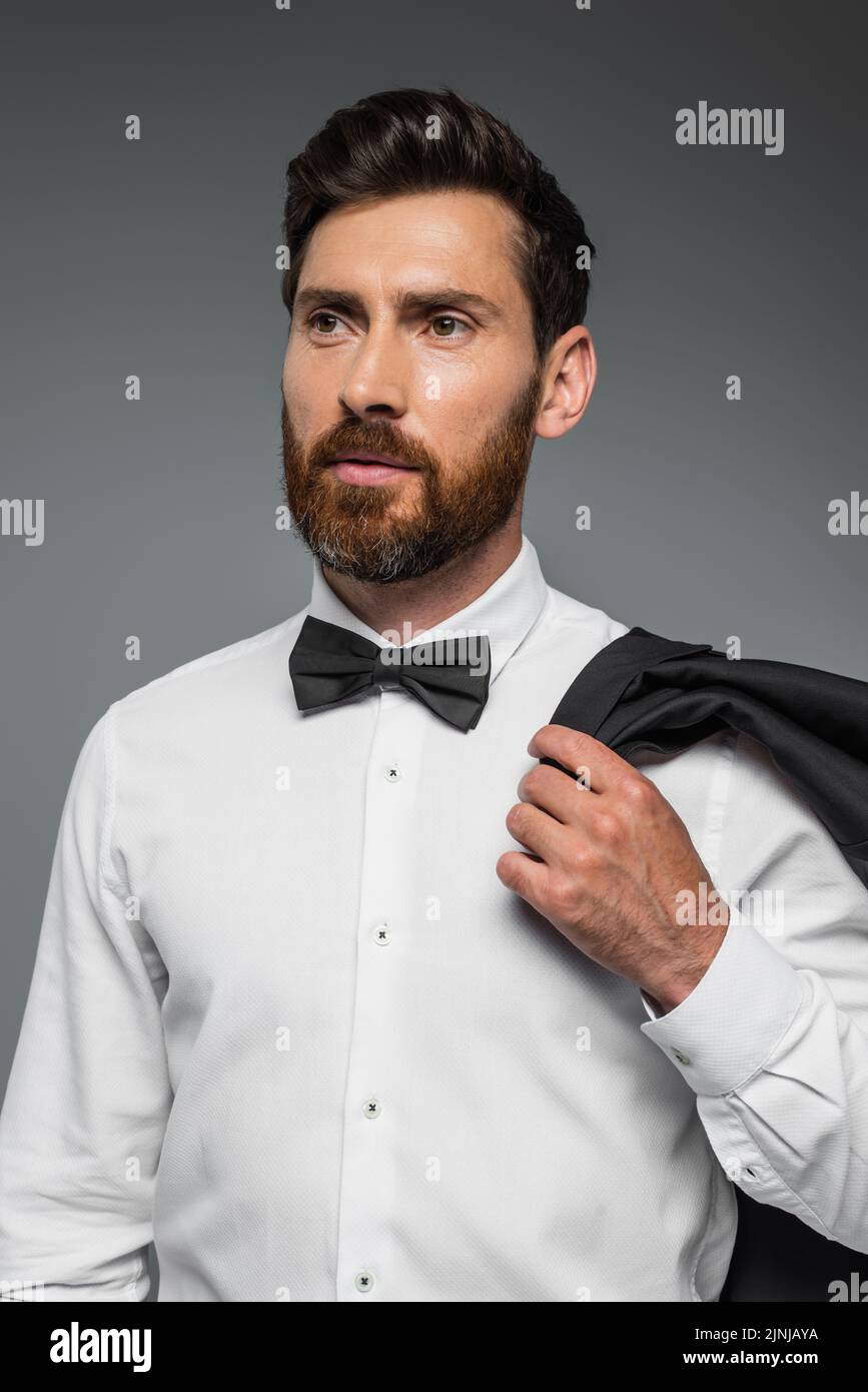 retrato de hombre barbudo en traje con lazo sujetando blazer aislado en gris, imagen de stock Foto de stock