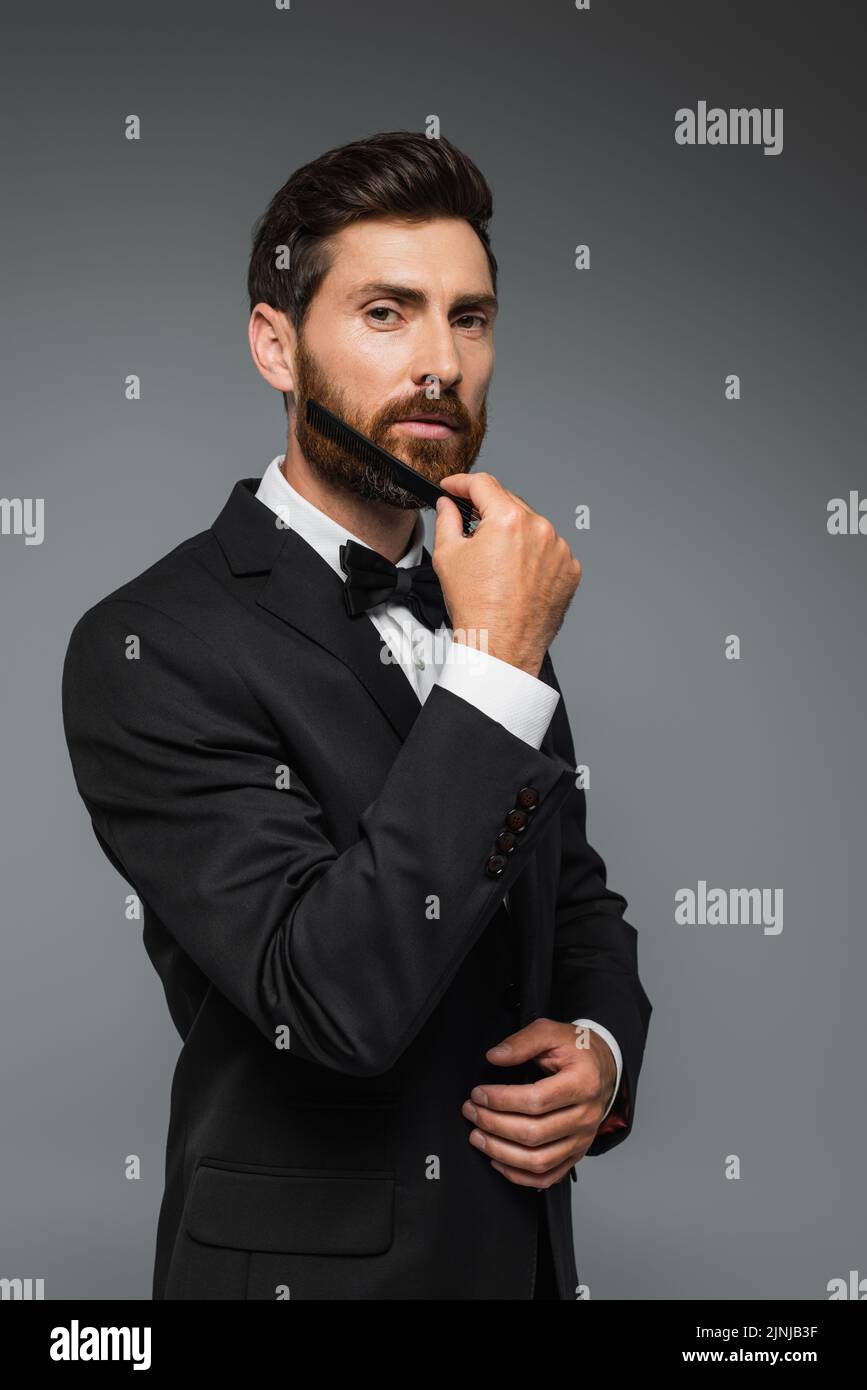 hombre con elegante traje de barba cepillando con peine aislado en gris, imagen de archivo Foto de stock