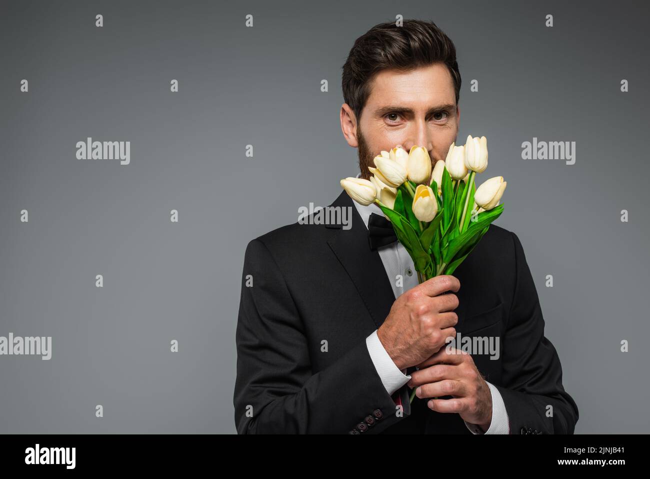 hombre barbudo en elegante tulipanes frescos con olor a esmoquin aislados en gris, imagen de archivo Foto de stock