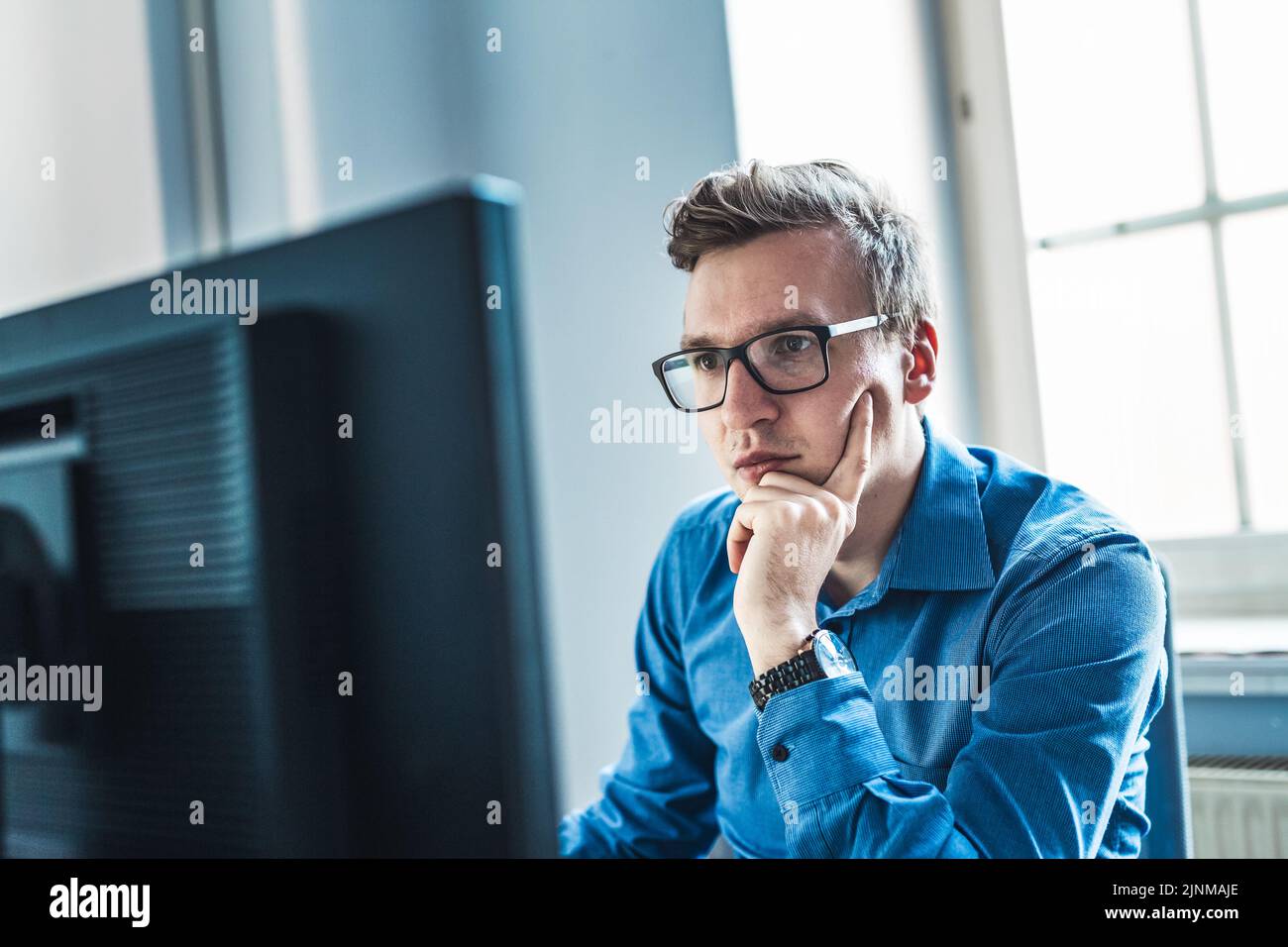 Guapo Joven hombre de negocios usando anteojos sentado en su mesa dentro de la oficina, mirando el informe en la pantalla de su computadora Foto de stock