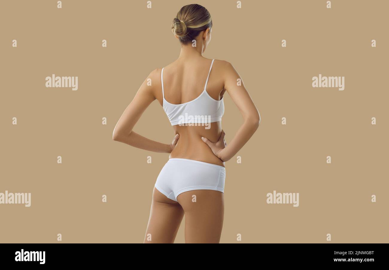 Vista posterior de modelo femenino joven y flaco posando en lencería blanca sobre fondo beige Foto de stock