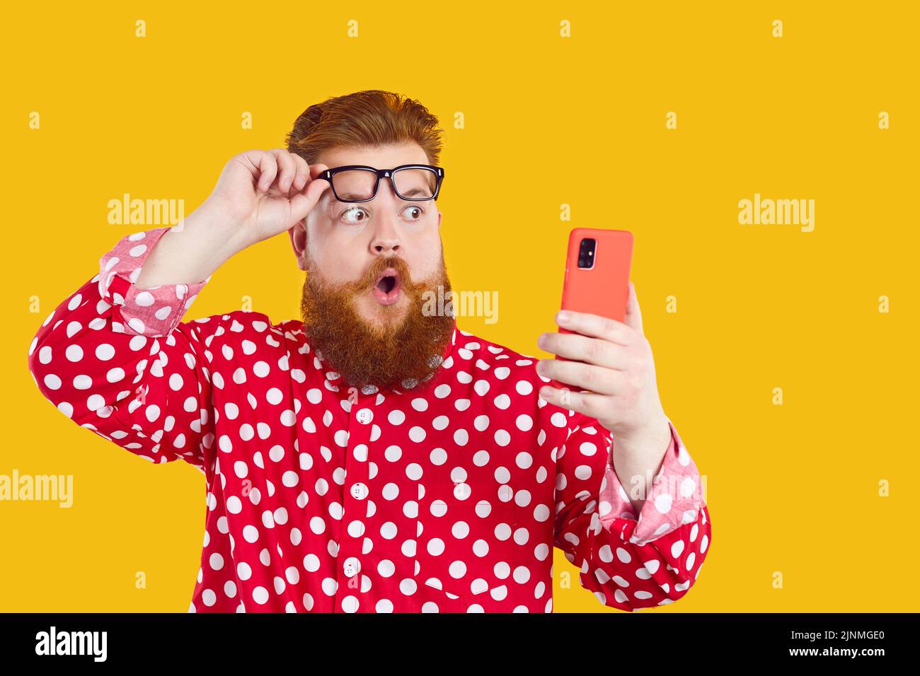 Gracioso hombre con expresión conmocionada mira la pantalla del teléfono móvil, levantando sus gafas. Foto de stock