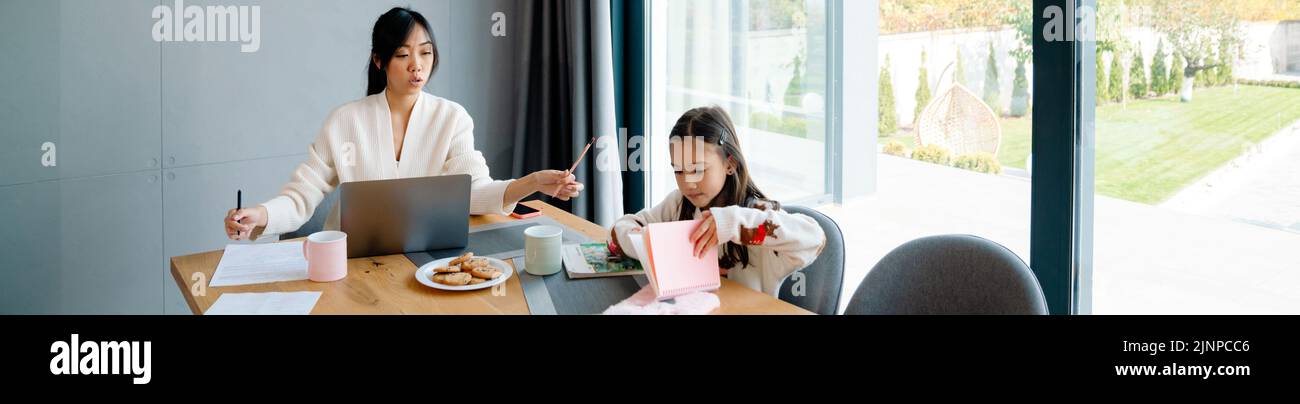 Mujer asiática que trabaja con el ordenador portátil mientras su hija hace los deberes en casa Foto de stock