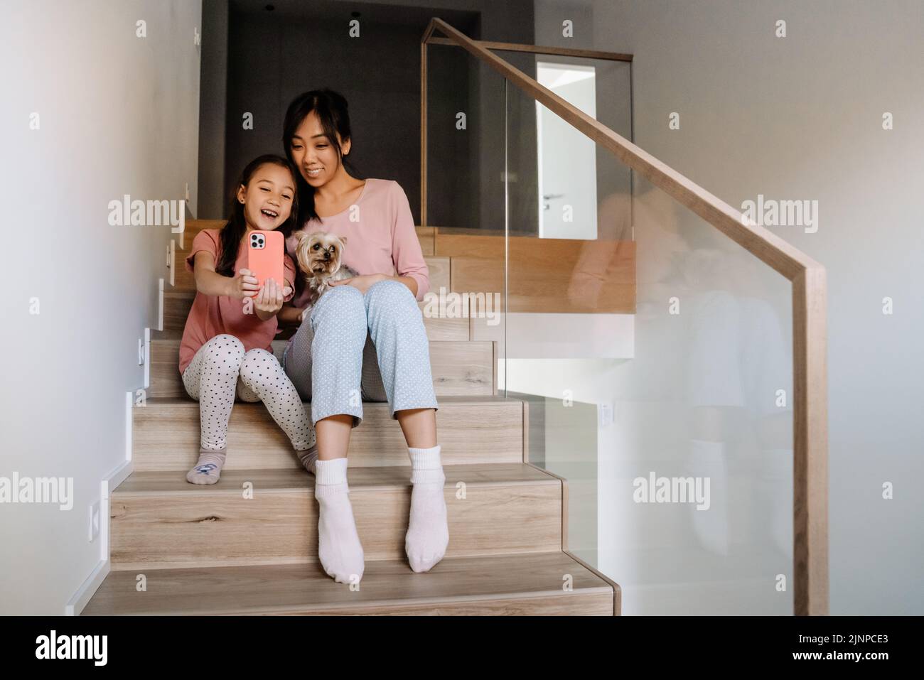 Madre asiática y su hija tomando selfie mientras se sientan con su perro en las escaleras en casa Foto de stock
