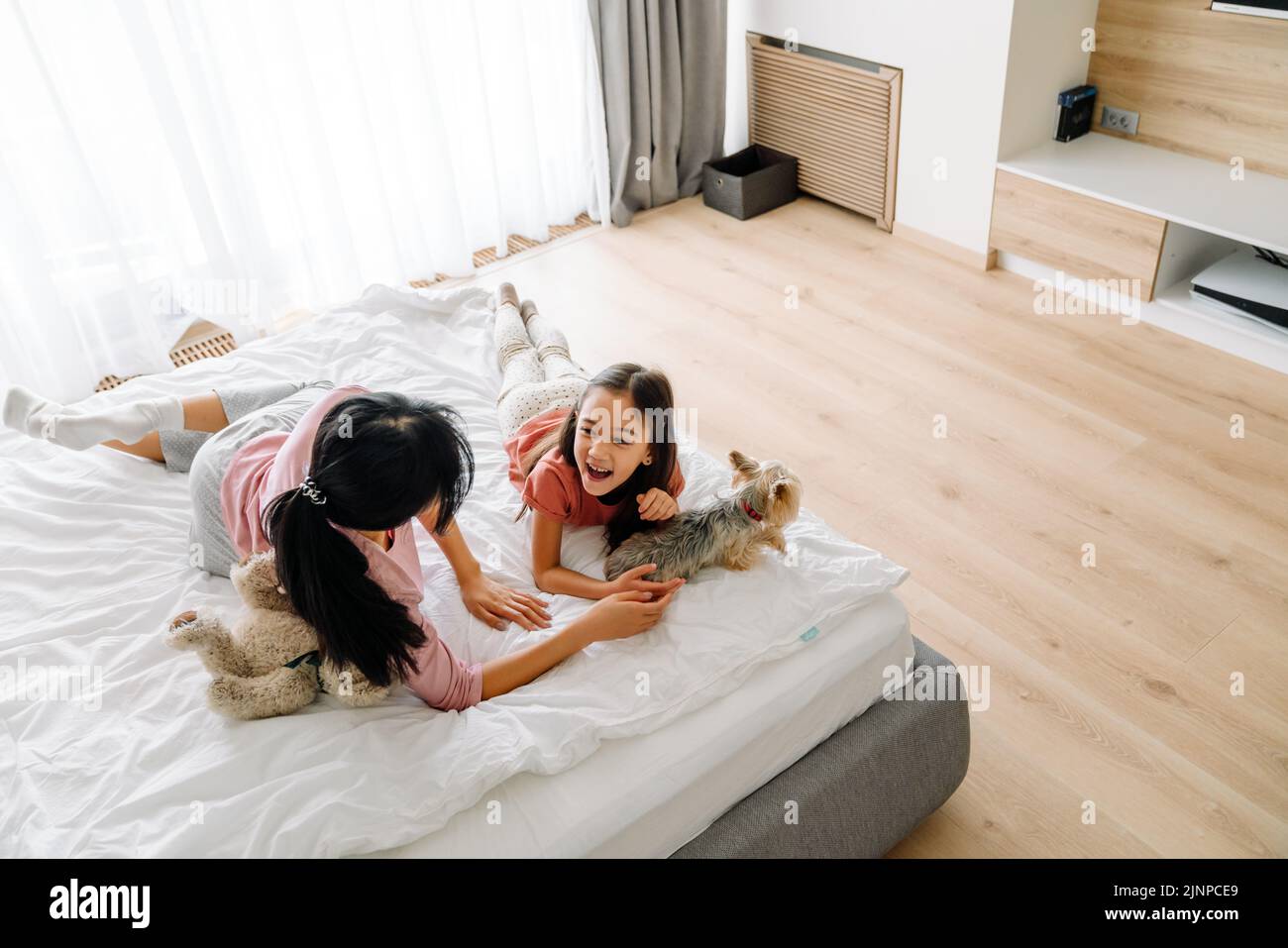 Madre asiática y su hija riendo mientras yacía en la cama con su perro en casa Foto de stock