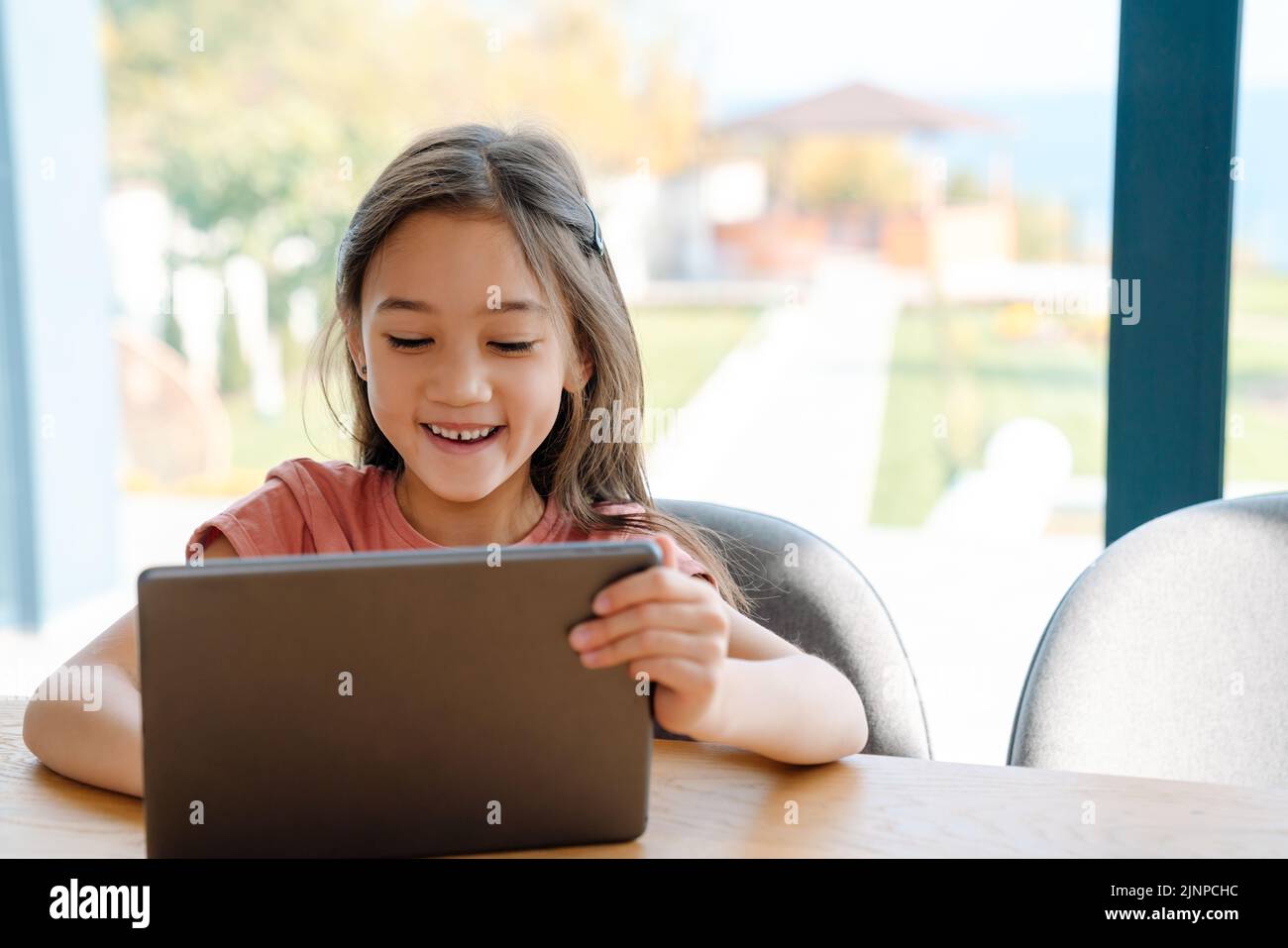 Chica asiática con risa y tableta de ordenador mientras se sienta en la mesa en casa Foto de stock